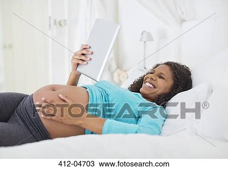 Stock Image - Pregnant woman using tablet computer on bed. Fotosearch