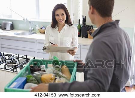 Stock Image - Woman signing for delivery in kitchen. Fotosearch