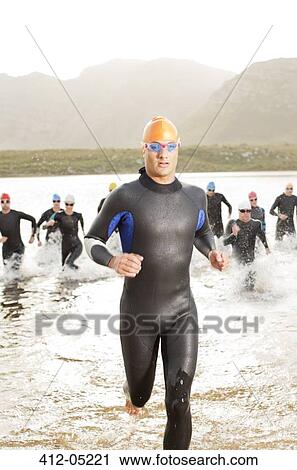 Stock Image - Triathletes in wetsuit running in waves. Fotosearch