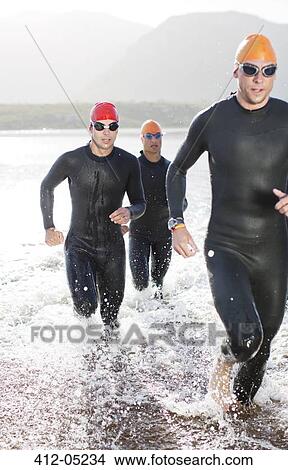 Picture - Triathletes in wetsuits running in waves. Fotosearch