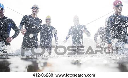 Stock Photo - Triathletes in wetsuits running in waves. Fotosearch