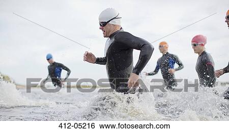 Stock Photograph - Triathletes in wetsuits running in waves. Fotosearch