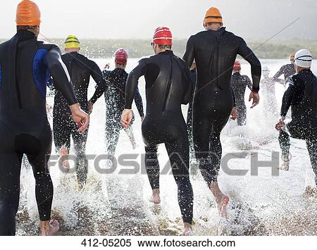 Stock Photography - Triathletes in wetsuits running in waves. Fotosearch