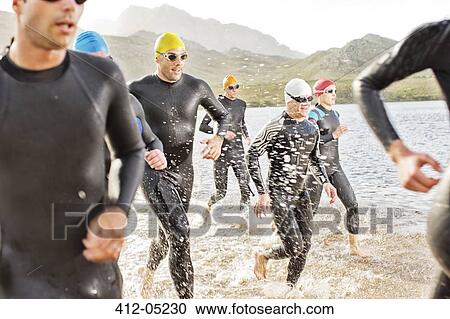 Stock Image - Triathletes in wetsuits walking in waves. Fotosearch