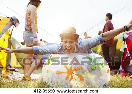 Stock Image - Portrait of playful man laying on inflatable chair outside tents at music festival. Fotosearch