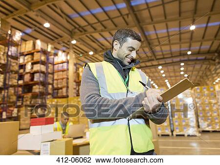 Stock Image - Worker writing on clipboard in warehouse. Fotosearch