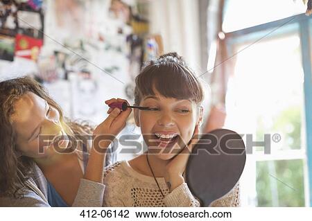 Stock Image - Woman applying makeup to friend's eyes in bedroom. Fotosearch