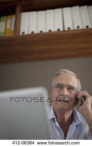 Picture - Older man talking on cell phone at desk. Fotosearch