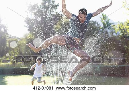 Stock Image - Father and son playing in sprinkler in backyard. Fotosearch