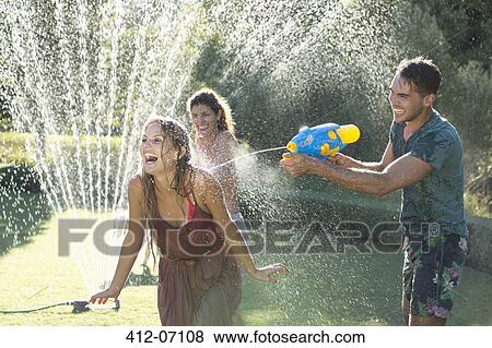Friends playing with water guns in sprinkler in backyard View Large Photo Image Stock Photo - Friends playing with water guns in sprinkler in backyard. Fotosearch