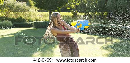 Stock Image - Girl playing with water gun in backyard. Fotosearch