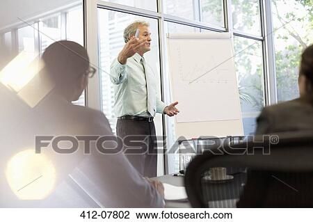 Businessman gesturing at flipchart in meeting View Large Photo Image Stock Image - Businessman gesturing at flipchart in meeting. Fotosearch