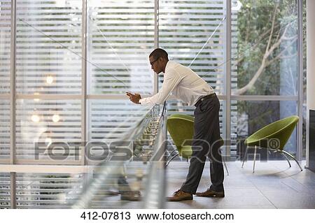 Stock Image - Businessman leaning on railing in office and text messaging with cell phone. Fotosearch