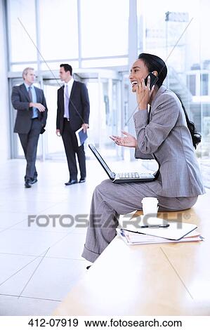 Stock Photo - Smiling businesswoman with laptop talking on cell phone in lobby. Fotosearch