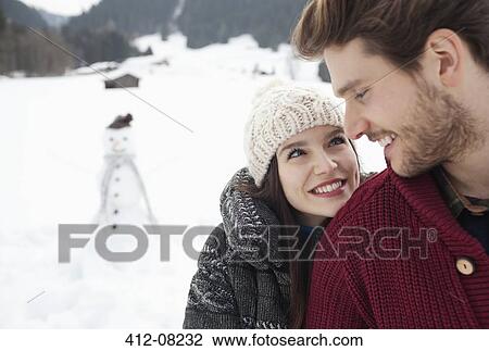 Stock Image - Close up of happy couple in snowy field with snowman. Fotosearch
