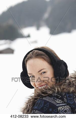 Stock Image - Close up portrait of woman wearing earmuffs in snowy field. Fotosearch
