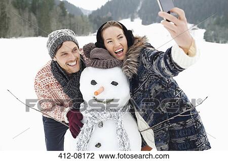 Stock Image - Enthusiastic couple taking self-portrait with snowman in snowy field. Fotosearch
