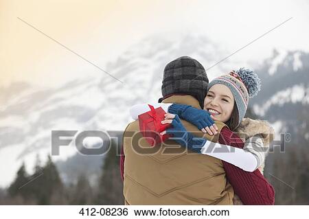 Stock Photograph - Happy woman holding gift and hugging man with mountains in background. Fotosearch