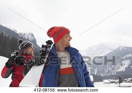 Stock Photography - Smiling men carrying skis at base of mountain. Fotosearch