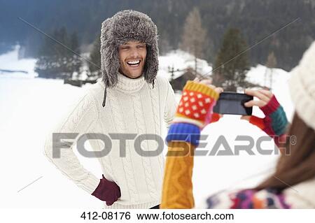 Stock Photograph - Woman photographing man in fur hat in snowy field. Fotosearch