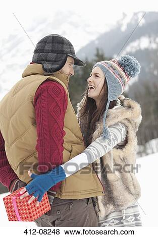 Stock Photo - Woman reaching for Christmas gift behind man's back in snowy field. Fotosearch