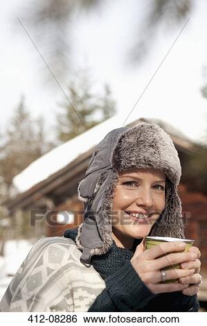 Stock Photograph - Close up portrait of woman in fur hat drinking coffee outside cabin. Fotosearch