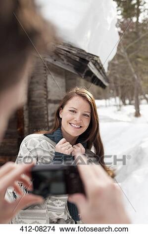 Picture - Man photographing smiling woman in snow outside cabin. Fotosearch