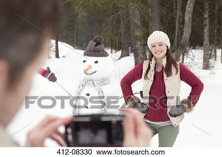 Stock Image - Man photographing smiling woman with hands on hips next to snowman in woods. Fotosearch