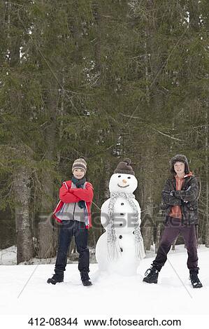Picture - Portrait of confident boys next to snowman in woods. Fotosearch