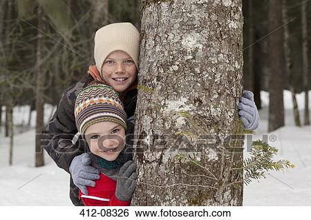 Stock Photograph - Portrait of happy boys behind tree trunk in snowy woods. Fotosearch
