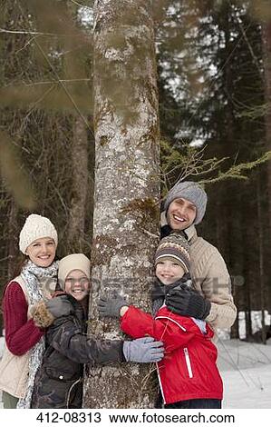 Stock Image - Portrait of happy family hugging tree trunk in snowy woods. Fotosearch