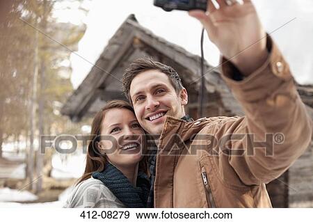 Stock Photo - Smiling couple taking self-portrait with digital camera outside snowy cabin. Fotosearch