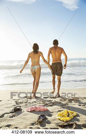 Stock Photo - Couple holding hands and walking toward ocean on beach. Fotosearch