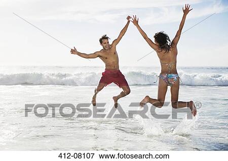 Stock Photo - Couple jumping with arms raised on beach. Fotosearch