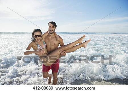 Stock Photo - Enthusiastic man carrying woman on beach. Fotosearch