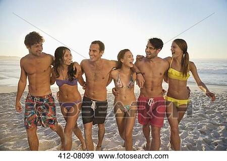 Stock Photo - Happy friends in bikinis and swim trunks walking on beach. Fotosearch
