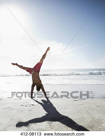 Stock Image - Man in swim trunks doing handstand on beach. Fotosearch