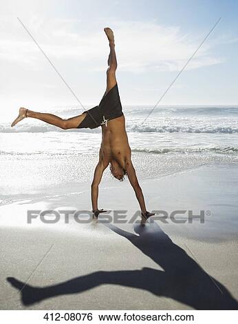 Stock Photograph - Man in swim trunks doing handstand on beach. Fotosearch