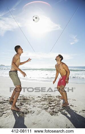 Stock Image - Men in swim trunks heading soccer ball on beach. Fotosearch
