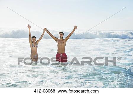 Stock Photo - Portrait of enthusiastic couple holding hands with arms raised in ocean. Fotosearch