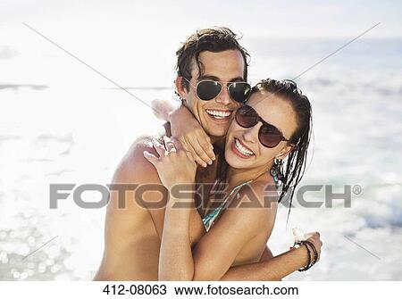 Stock Image - Portrait of enthusiastic couple in sunglasses hugging on beach. Fotosearch