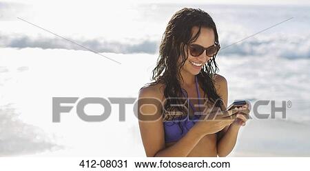 Stock Image - Smiling woman text messaging with cell phone on beach. Fotosearch