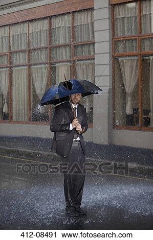 Stock Image - Businessman standing under broken umbrella in rainy street. Fotosearch