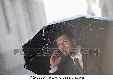 Picture - Businessman talking on cell phone under umbrella in rain. Fotosearch