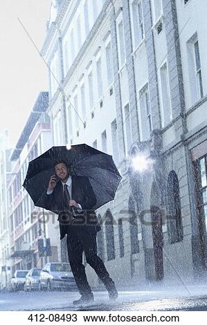 Stock Image - Businessman talking on cell phone under umbrella in rainy street. Fotosearch
