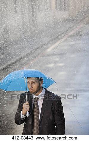 Stock Image - Businessman under tiny umbrella in rainy street. Fotosearch