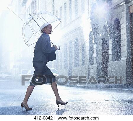 Stock Image - Happy businesswoman with umbrella walking across rainy street. Fotosearch