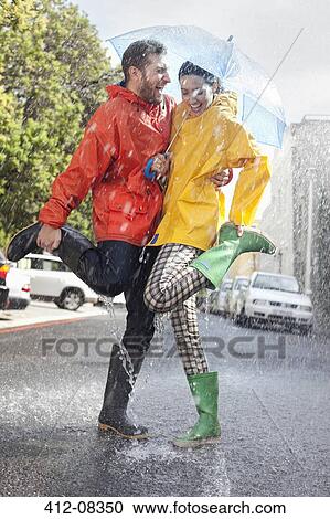Stock Image - Happy couple in wellingtons splashing in rainy street. Fotosearch