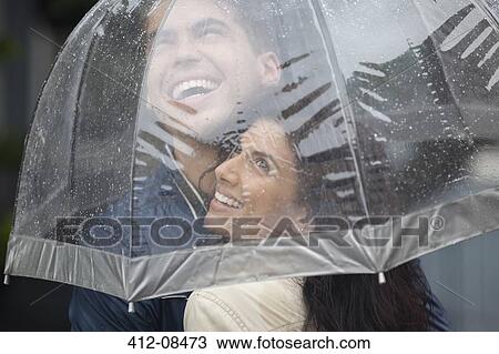 Stock Image - Happy couple under umbrella looking up at rain. Fotosearch