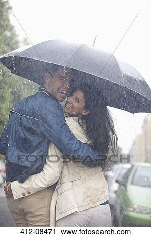 Stock Image - Portrait of happy couple hugging under umbrella in rain. Fotosearch